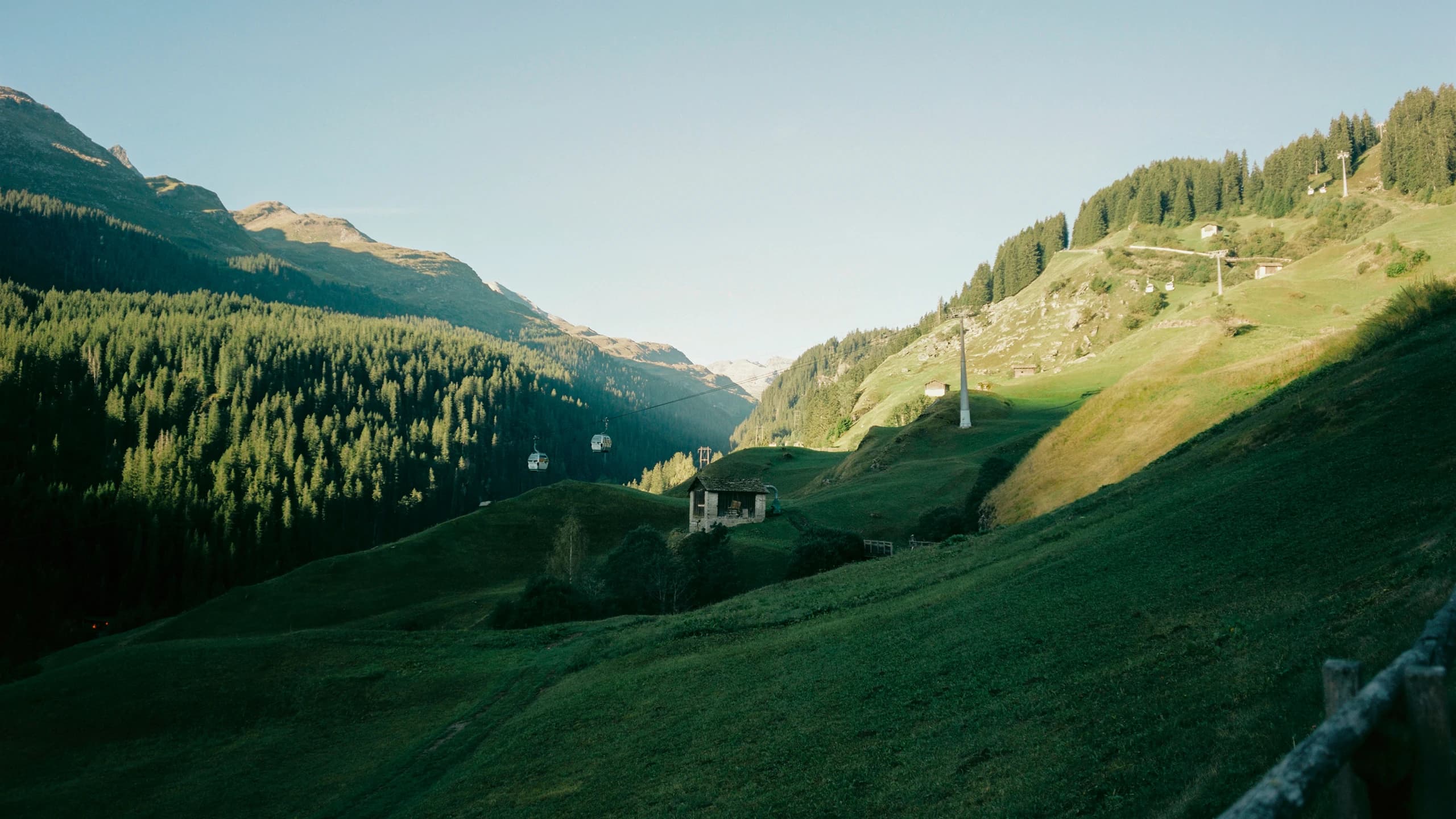 A green mountain valley with a forest, a farmhouse, and a gondola lift in the evening light