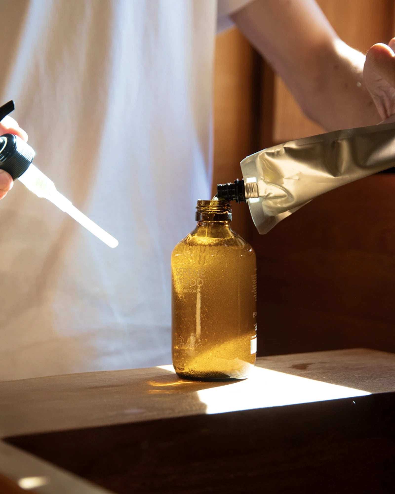 A person pours liquid soap from a refill pouch into a brown glass bottle